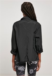 Person med långt flätat hår som bär en lös svart jacka med uppvikt ärmar och svarta shorts med blommönster, vänd bortåt mot en ljus bakgrund.