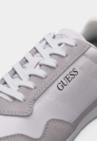 Baskets blanches et gris clair avec lacets blancs et logo "GUESS" sur le côté, présentant des détails en suède cousu et en tissu.