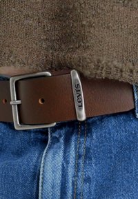 Brauner Ledergürtel mit einer silbernen Metallspange, die das "Levi's"-Logo präsentiert, getragen über blauen Jeans mit hellblauen und goldenen Nähten.