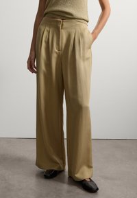 Helle khakifarbene weite Beine mit Falten, glatter Textur, hoher Taille, seitlichen Taschen und lässiger Passform. Getragen mit schwarzen spitzen Schuhen.