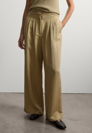 Pantaloni a gamba larga color khaki chiaro con pieghe, texture liscia, vita alta, tasche laterali e vestibilità rilassata. Indossati con scarpe nere a punta.