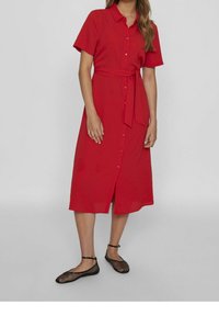 Robe rouge à manches courtes boutonnée avec une ceinture à la taille, longueur jusqu'aux genoux, en tissu lisse, avec une fente à l'avant, associée à des ballerines noires en maille.