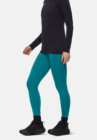Leggings turchesi con un design aderente, caratterizzati da cuciture a contrasto e una tasca laterale, abbinati a una maglietta nera a maniche lunghe e scarpe nere.