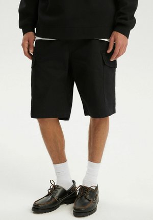 Osoba ubrana w czarne krótkie spodnie cargo, białe skarpetki i czarne sznurowane buty, stojąca na jednolitym białym tle.