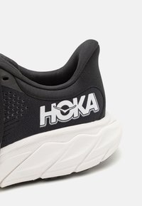 Chaussure de sport noire avec tige en mesh, col rembourré, branding HOKA blanc et semelle blanche texturée avec des motifs de traction distincts.
