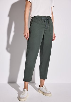 Personne portant un pantalon vert foncé à cordon, des baskets blanches et un t-shirt blanc à manches courtes, debout devant un fond uni.