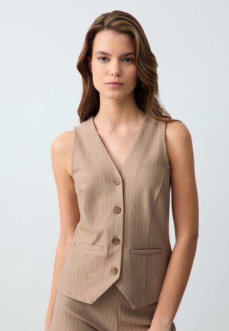 Gilet beige con righe bianche verticali, design a V, cinque bottoni marroni e tasche laterali. Tessuto liscio con una silhouette su misura.