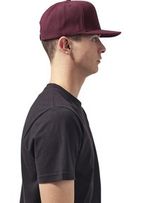 Burgundy színű baseballsapka strukturált kialakítással és ívelt karimával, amelyet egy fekete pólót viselő személy hord, oldalról nézve.