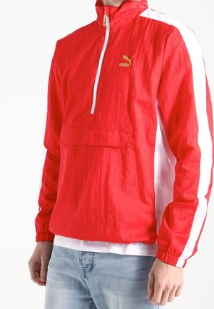 Punainen pitkähihaisen Puma-pullover, jossa on valkoiset raidat hihoissa ja edessä vetoketju, puettu valkoisen paidan päälle ja yhdistetty vaaleansinisiin farkkuihin.