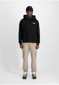 Sudadera de algodón negra con bolsillo, pantalones cargo beige con bolsillos laterales y zapatillas negras con acentos blancos. Diseño simple, ajuste casual.
