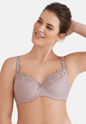 Frau lächelt und trägt einen hell mauvefarbenen BH mit Spitzenbesatz und dekorativen Mustern auf den Cups und Trägern, posiert mit einem erhobenen Arm.