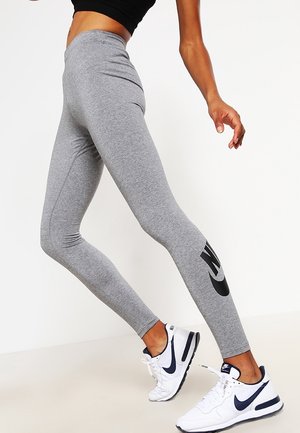 Leggings gris en tissu jersey, dotés d'une taille haute et d'un grand logo Nike noir sur la jambe inférieure, associés à des chaussures de sport blanches.