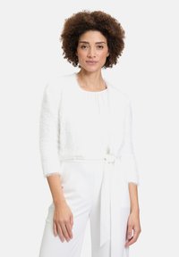 Biała, puszysta krótka kurtka z rękawami 3/4, otwartym przodem i teksturowanym wykończeniem. Nosi ją się na białej bluzce i spodniach z paskiem.