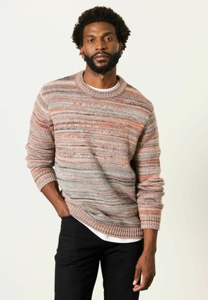 Mann mit Bart, der einen mehrfarbig gestreiften Strickpullover mit weißem Unterhemd und schwarzen Hosen trägt, steht vor einem einfachen hellen Hintergrund.
