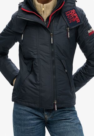 Persoon draagt een marineblauw Superdry jas met rits en rode accenten over een beige gebreide trui en blauwe spijkerbroek, poserend met handen in de jaszakken.