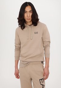 Beige hettegenser med en frontlomme og snor, med et logo på brystet. Kombinert med matchende bukser som har et teksturert mønster og logo.