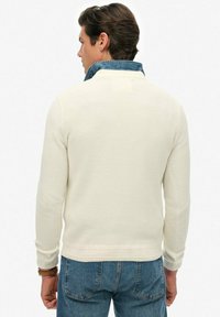 Mężczyzna z brązowymi włosami, ubrany w kremowy sweter dziany na koszulę z kołnierzykiem z denimu oraz niebieskie dżinsy, odwrócony plecami do aparatu.