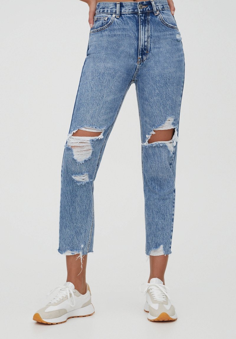 Jeans dżinsowe w kolorze niebieskim z wysokim stanem, z przetarciami i dużymi dziurami na kolanach, o prostym kroju nogawki i surowym wykończeniu. Noszone z białymi trampkami.