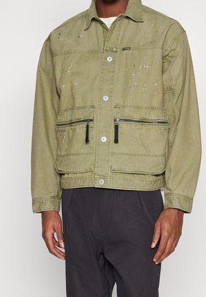Giubbotto di jeans verde con tasche frontali, chiusure a zip argentate e macchie di vernice. Presenta un'apertura con bottoni e un design del colletto.