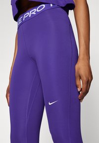 Fialové sportovní legíny s přiléhavým střihem, hladkou texturou a výrazným bílým logem Nike na levém stehně. Pásek v pase nese branding "NIKE PRO".