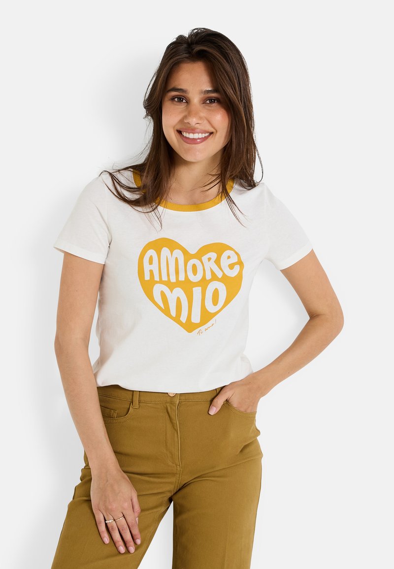 Camisa de algodón blanca con cuello amarillo y gráfico en forma de corazón con la frase "AMORE MIO" en letras negritas. Combinada con pantalones de color mostaza.