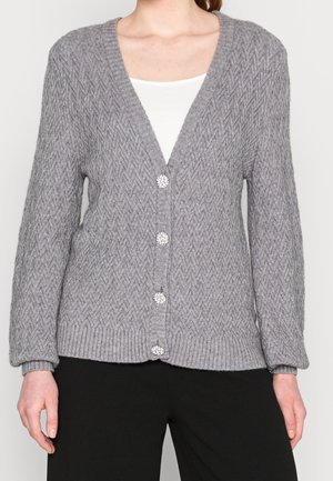 Frau trägt eine graue gestrickte Strickjacke mit gemustertem Muster und perlenähnlichen Knöpfen über einem weißen Oberteil und schwarzen Hosen.