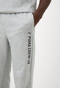 Pelēki kokvilnas sporta bikses ar sānu kabatu un melnu drukātu tekstu "PUMA CREW '48" ar mazu logo uz kājas. Gluda tekstūra.