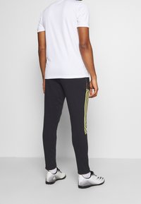 Svarta träningsbyxor med gula sidostråk, ihopparat med en vit t-shirt med korta ärmar. Har en figursydd design och en slät textur.