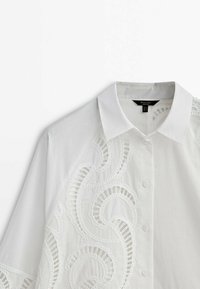 Chemise blanche avec un col classique, présentant une broderie blanche délicate sur un côté et des boutons sur le devant. Tissu doux et lisse.