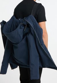 Veste bleu marine en matière résistante à l'eau, dotée d'une capuche amovible et d'une fermeture éclair, portée par-dessus une chemise noire.