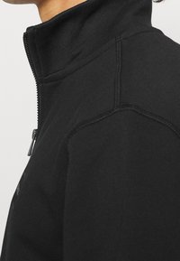 Vista lateral de una persona que lleva una chaqueta negra de cremallera con cuello alto y detalles de costura visibles en el hombro y la manga.