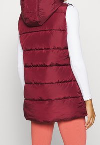 Burgundowa kamizelka puchowa bez rękawów z kapturem, z pionowymi przeszyciami, gładką teksturą i zwężanym krojem, zestawiona z jasnokolorowymi długimi rękawami.