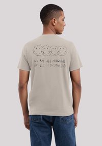 Light beige cotton t-shirt featuring a playful graphic of four smiley faces and text reading "WE ARE ALL ORIGINAL SMILEY ORIGINALS" on the back.