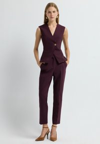 Blazer violaceo senza maniche con scollatura a V profonda e bottone dorato, abbinato a pantaloni su misura dello stesso colore e scarpe con tacco alte marroni.