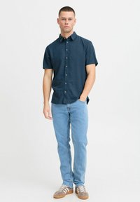 Chemise à manches courtes bleu marine à boutons, associée à un jean bleu clair et des baskets en daim marron avec des accents blancs. Design sobre, coupe décontractée.