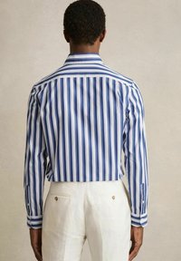 Chemise rayée bleue et blanche, à manches longues, avec des poignets boutonnés et un col classique, associée à un pantalon couleur crème avec des poches arrière.