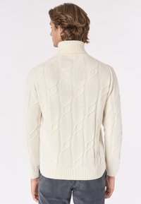 Uomo con capelli castano chiaro che indossa un maglione a collo alto di colore crema con motivo a treccia e pantaloni grigio scuro, mostrato da dietro.