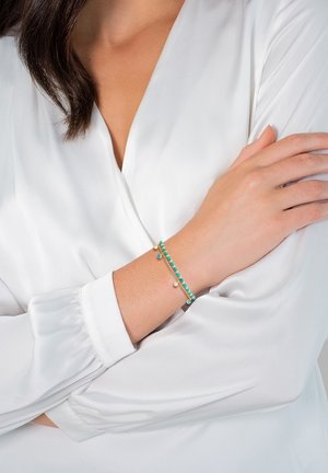 Guld-tone armbånd med turkise og blå perler, med et delikat design og cirkulære accenter, båret på et håndled mod en hvid ærme.