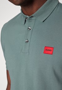 Homme portant un polo vert à trois boutons avec un patch logo carré rouge indiquant HUGO sur la poitrine.