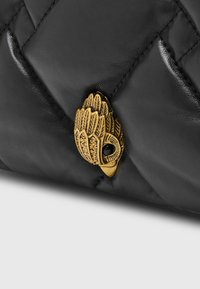 Zlatý klip ve tvaru sovy na černém prošívaném koženém materiálu, pravděpodobně součást tašky nebo doplňku.