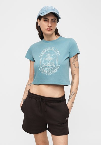 Νεαρή γυναίκα με σκουλαρίκια και τατουάζ, φοράει μπλε κοντομάνικο μπλουζάκι με γραφικό συντριβάνι, μαύρο σορτς με κορδόνι και ανοιχτό μπλε καπέλο.