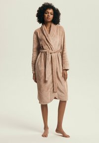 Woman standing barefoot wearing a knee-length, light brown plush bathrobe tied at the waist against a plain background.