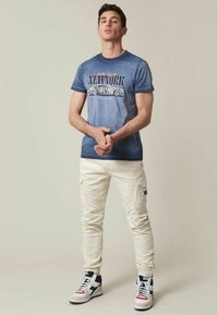 T-shirt dégradé bleu avec un motif "NEW YORK" ; pantalon cargo blanc ; sneakers blancs avec des accents bleu marine et rouge ; posé contre un fond neutre.