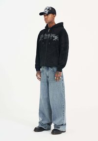 Black zip-up hoodie with textured pattern and silver graphic. Light blue wide-leg jeans. Black cap with dragon design. Black shoes.