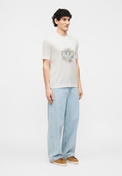 Νέος άνδρας που στέκεται, φοράει ανοιχτό μπεζ μπλουζάκι με γραφικά, φαρδιά ανοιχτά μπλε τζιν και καφέ σουέντ αθλητικά παπούτσια, μπρο στά σε απλό λευκό φόντο.