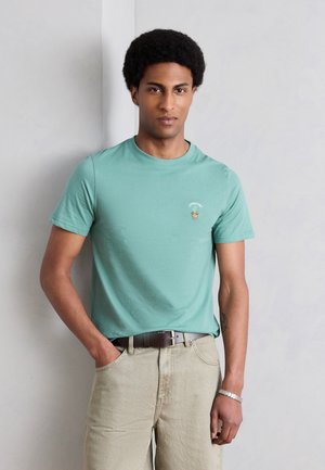 Mężczyzna z fryzurą afro, ubrany w miętową koszulkę z małym wzorem ananasa i beżowe spodnie, stoi przy szarej ścianie.