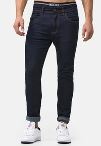 Ciemnoniebieskie obcisłe dżinsy z denimu z zapięciem na guzik, kontrastującym przeszyciem i podwiniętymi mankietami, zestawione z białymi trampkami.