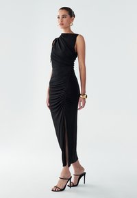 Kvinde i en ærmeløs sort rynket kjole med høj slids, sorte højhælede sandaler, guld hoop øreringe og et guld manchettearmbånd, der står.