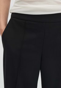 Pantalon noir sur mesure avec une texture lisse, doté de poches latérales et de plis subtils. La ceinture est élastiquée pour plus de confort.