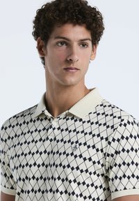 Poloshirt in Creme mit einem Rautenmuster in Marineblau. Verfügt über einen klassischen Kragen, eine Zweiknopf-Leiste und ein kleines Logo-Detail auf der Brust.